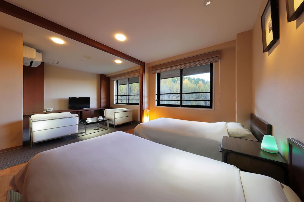 A hotel room with two beds, a desk, a chair, a TV, and a window with a view of trees.