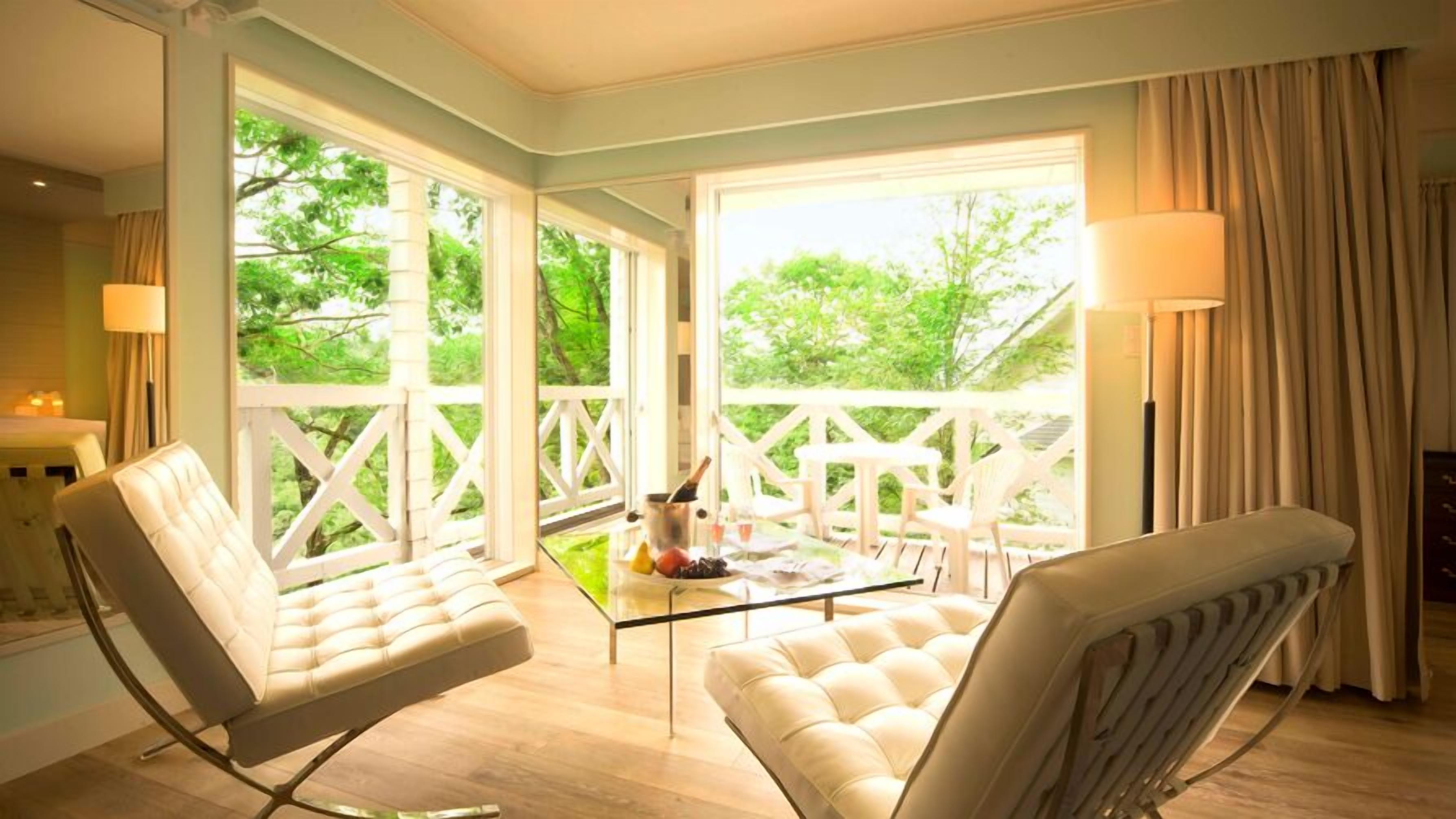 A room with two cushioned chairs, a glass table with drinks, and large windows with a view of greenery.