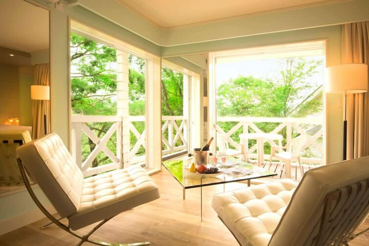 A room with two cushioned chairs, a glass table with drinks, and large windows with a view of greenery.