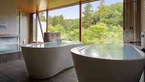 A spa area with two large tubs under a wooden ceiling, a stone wall, and a view of a forest. - Hotel Ryu Resort and Spa (Takayama)