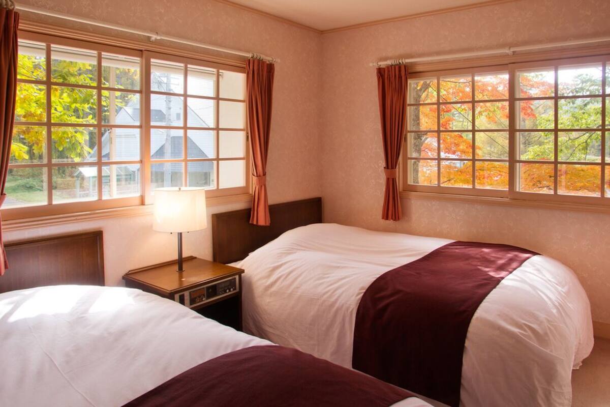 A hotel room with two beds, a nightstand, a lamp, and windows with curtains.
