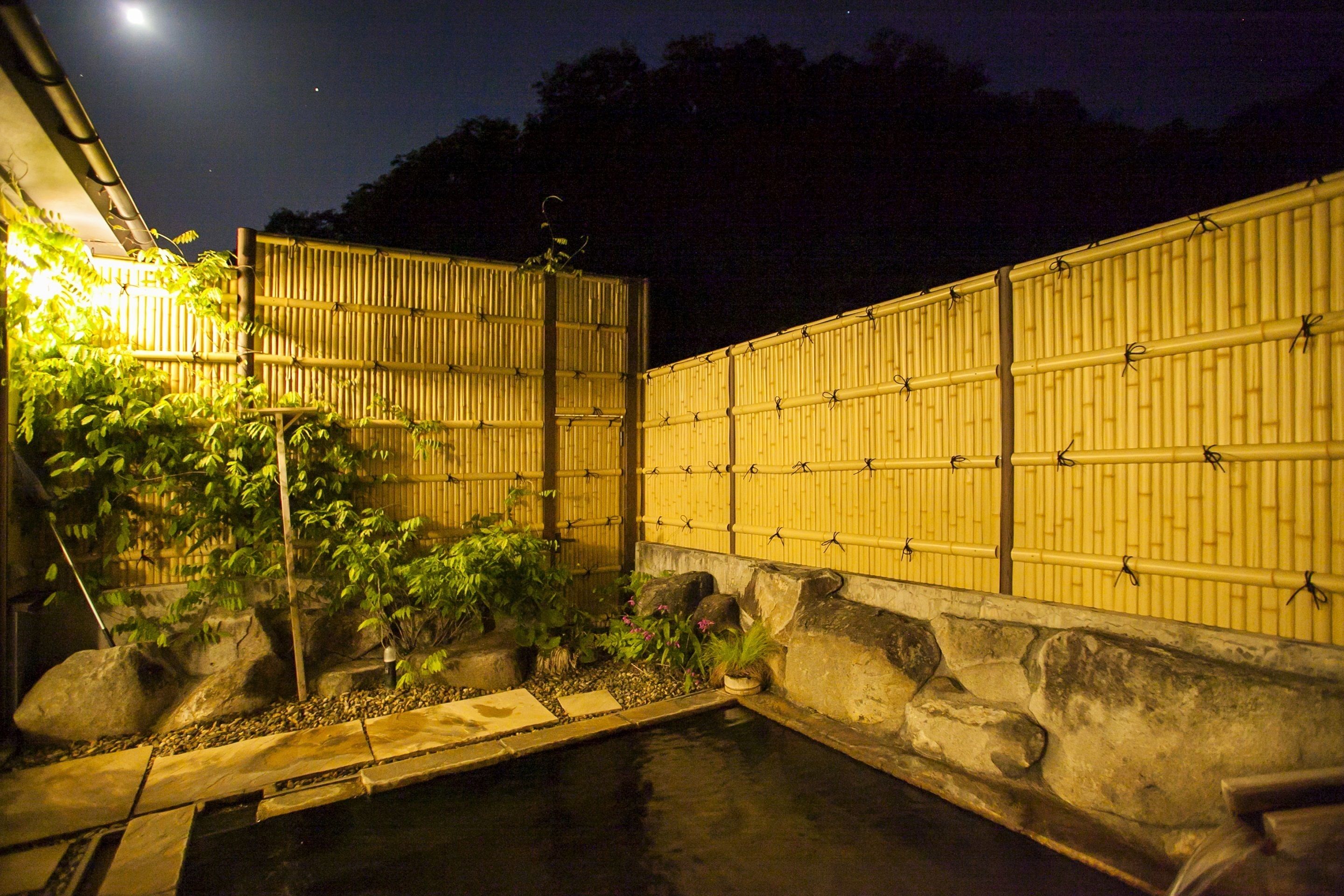 A traditional Japanese garden at night with a koi pond, bamboo fence, and illuminated plants.