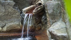 A small waterfall in a natural setting with rocks and a bamboo fence in the background.