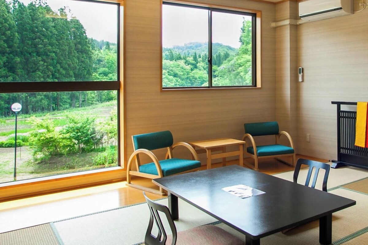Una habitación con suelo de tatami, una mesita baja, dos sillas y vistas a espacios verdes.