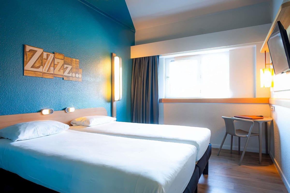 Een hotelkamer met twee bedden, een bureau, een stoel, een raam met gordijnen en een aan de muur bevestigde televisie.
