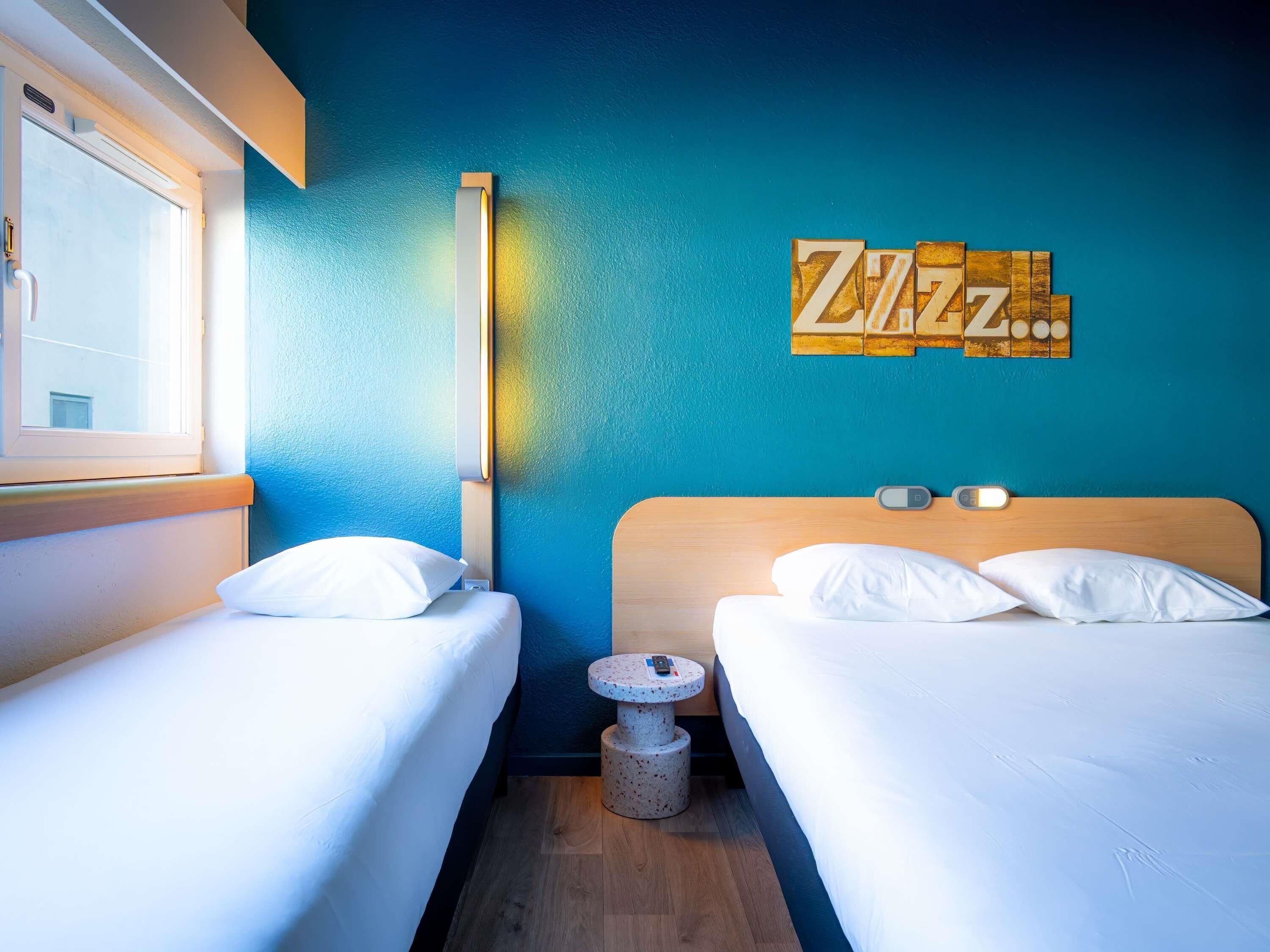 A hotel room with two beds, a small round table, a wall clock, and a window.