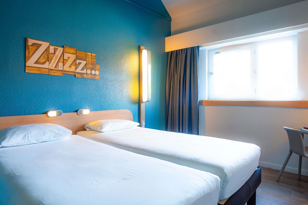 A hotel room with two beds, a desk, a window with curtains, and a wall with a wooden sign.