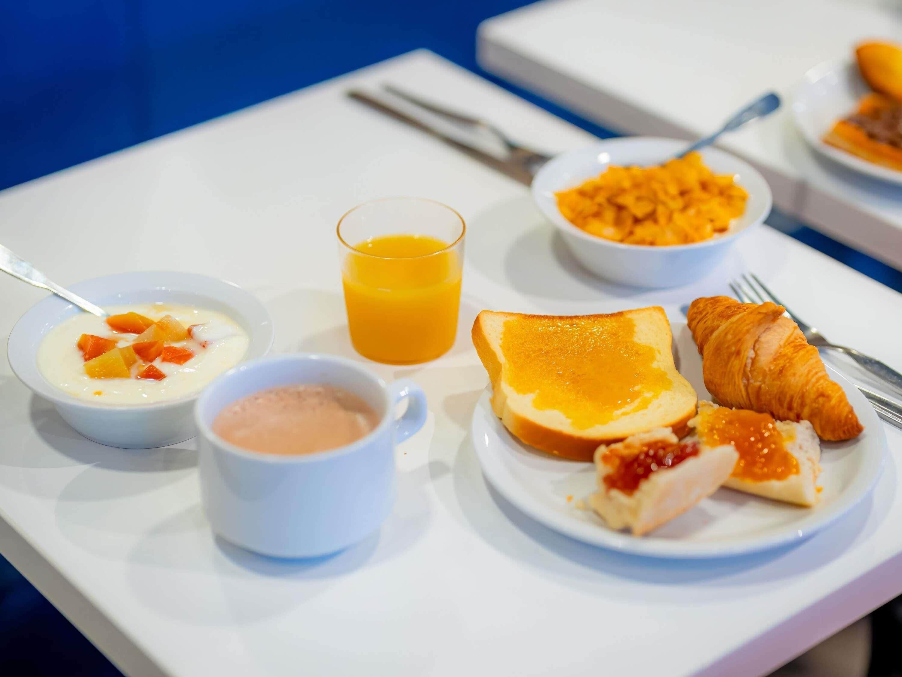 A breakfast setting with a plate of croissant, toast, and fruit, a bowl of yogurt with fruit, a glass of orange juice, and a small cup of coffee.