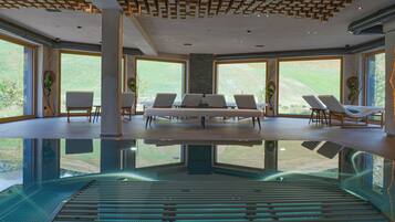 Uma área de piscina interior moderna com paredes de vidro transparente, cadeiras de descanso e um design de teto singular.