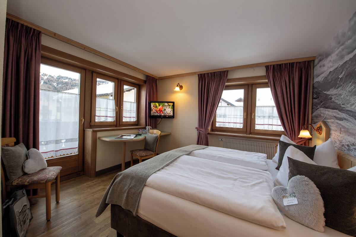 Una habitación de hotel con una cama grande, un escritorio con silla, televisión y vistas a las montañas desde las ventanas.