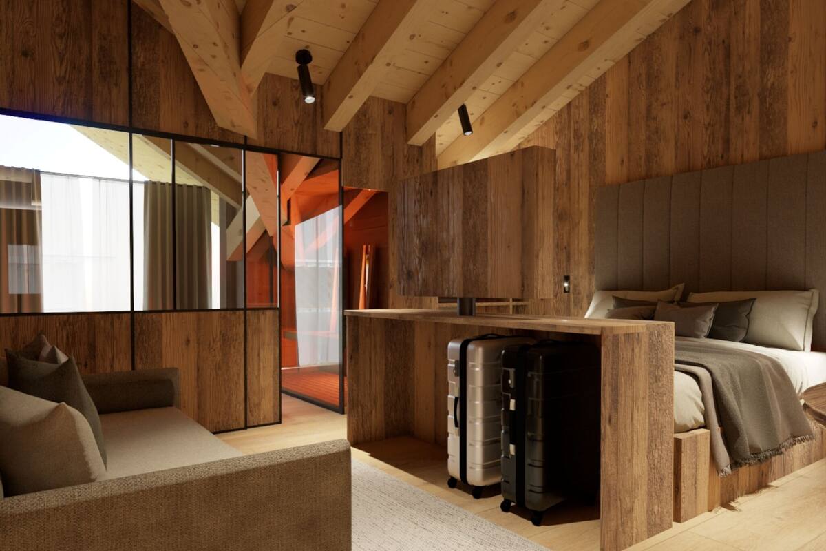 Una habitación moderna y rústica con techo de madera, una cama con almohadas, un sofá, un escritorio con una silla y un ventanal grande.