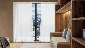 A cozy living room with a large window, wooden beams, a sofa, and a bookshelf. - Al Nin Hotel&Spa (Livigno)