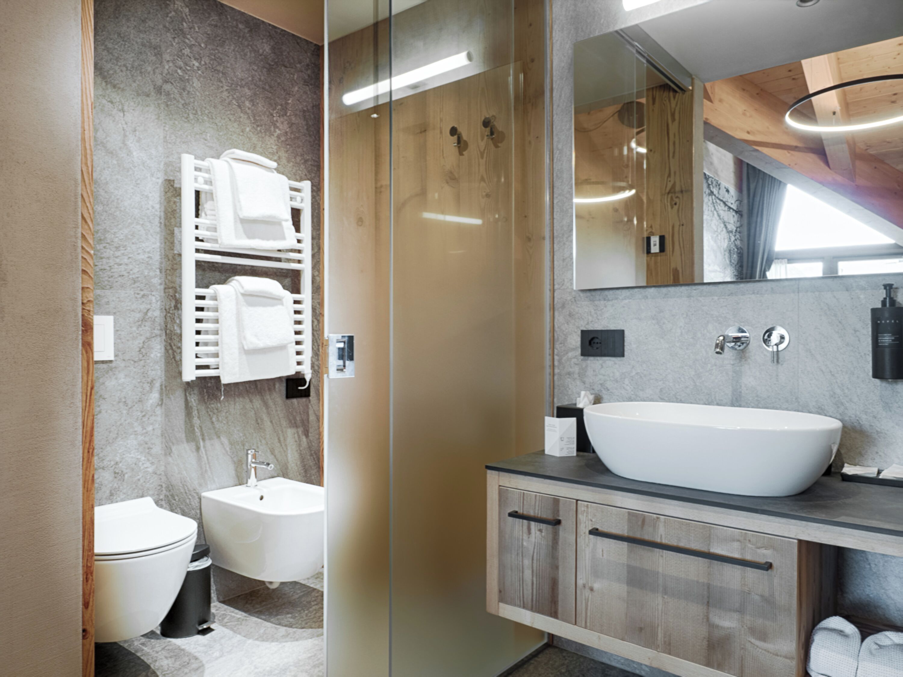 Un baño moderno con un lavamanos grande blanco, un bidé y un inodoro. Hay un mueble de tocador de madera con un espejo sobre el lavamanos.
