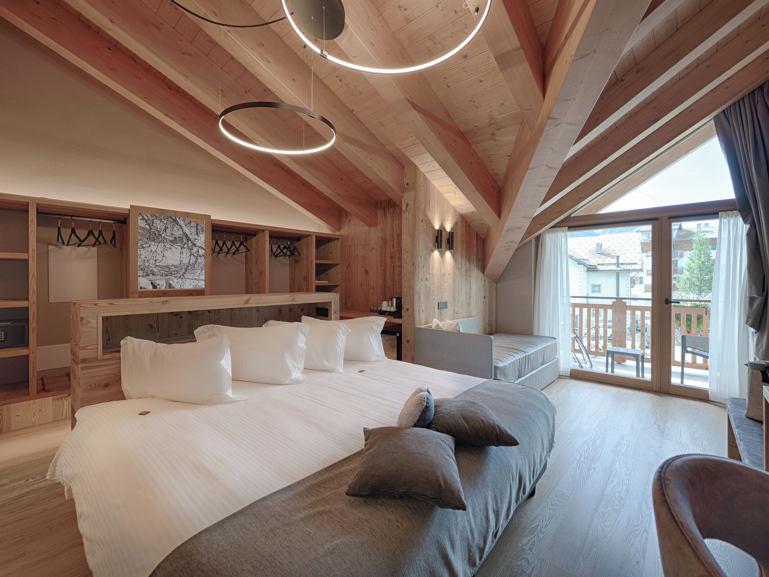 A bedroom with a large bed, a sofa, a wooden headboard, a painting, and a balcony with a view of buildings.