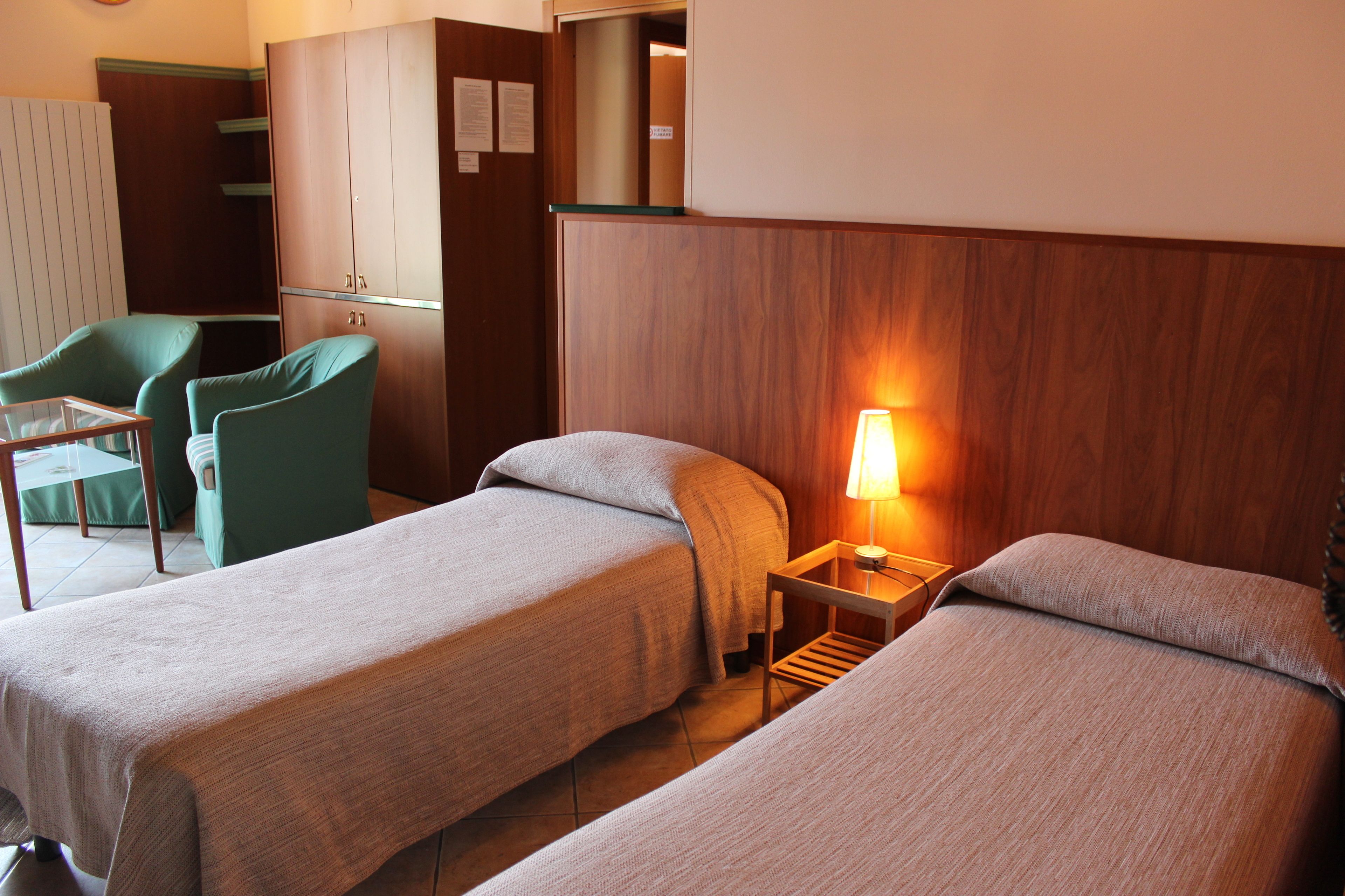 A hotel room with two single beds, a small table with a lamp, a green chair, and a wardrobe.