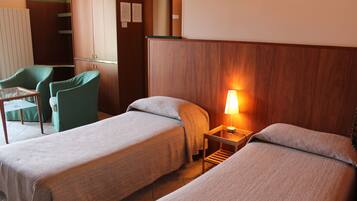 A hotel room with two single beds, a small table with a lamp, a green chair, and a wardrobe.