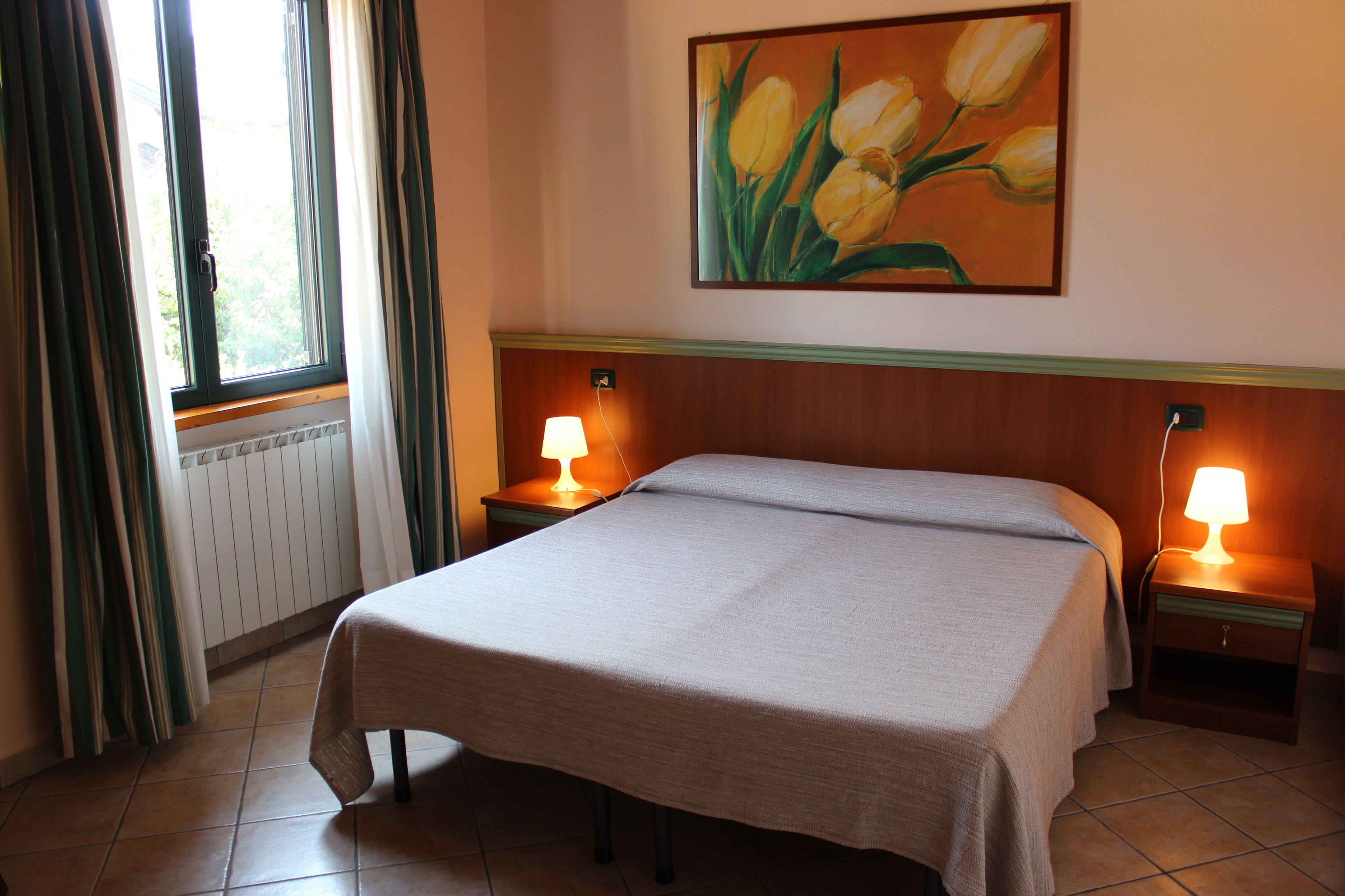 Une chambre d’hôtel avec un lit, deux tables de chevet avec des lampes, une fenêtre avec des rideaux et un tableau représentant des tulipes accroché au mur.