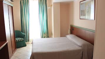 A hotel room with a bed, a green armchair, a window with striped curtains, and a painting on the wall.