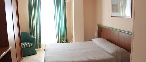 A hotel room with a bed, a green armchair, a window with striped curtains, and a painting on the wall.