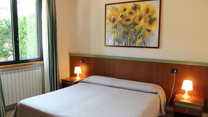 A neatly made bed with a light-colored bedspread, two bedside lamps, a painting of sunflowers on the wall, and a window with curtains. - B&B Villa Giglio (Arsago Seprio)