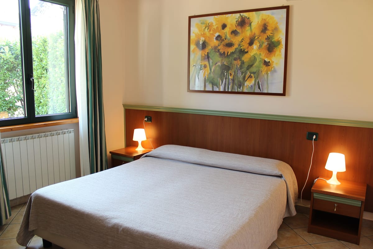 Un lit bien fait, recouvert d’une couvre-lit de couleur claire, deux lampes de chevet, un tableau représentant des tournesols accroché au mur et une fenêtre avec des rideaux.