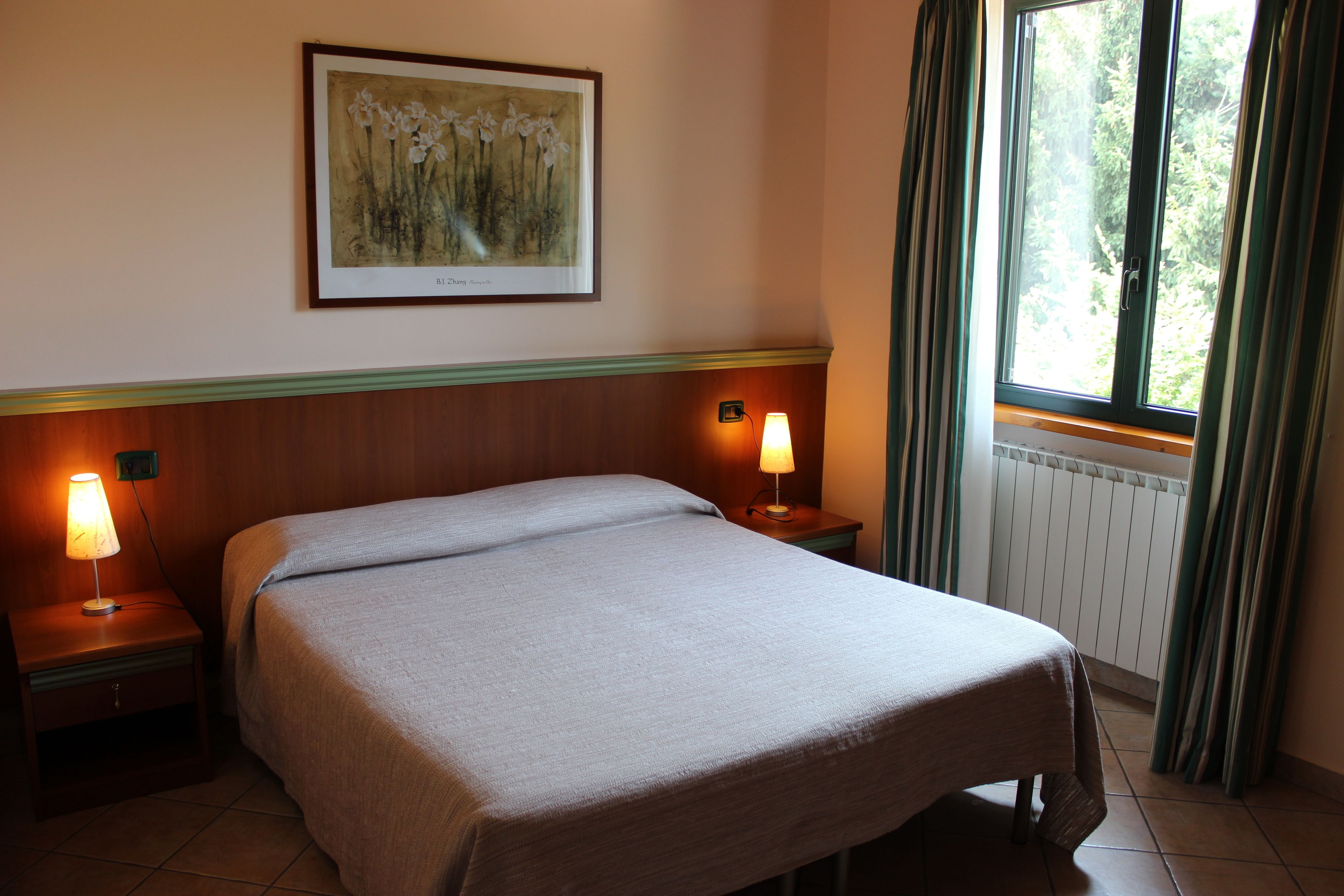 A hotel room with a bed, two bedside lamps, a framed artwork on the wall, a window with curtains, and a radiator.