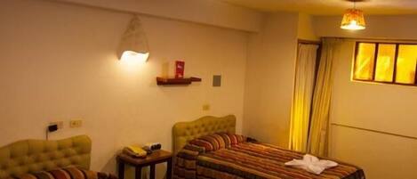 A hotel room with two beds, a small table, a wall-mounted shelf, and a window with curtains.