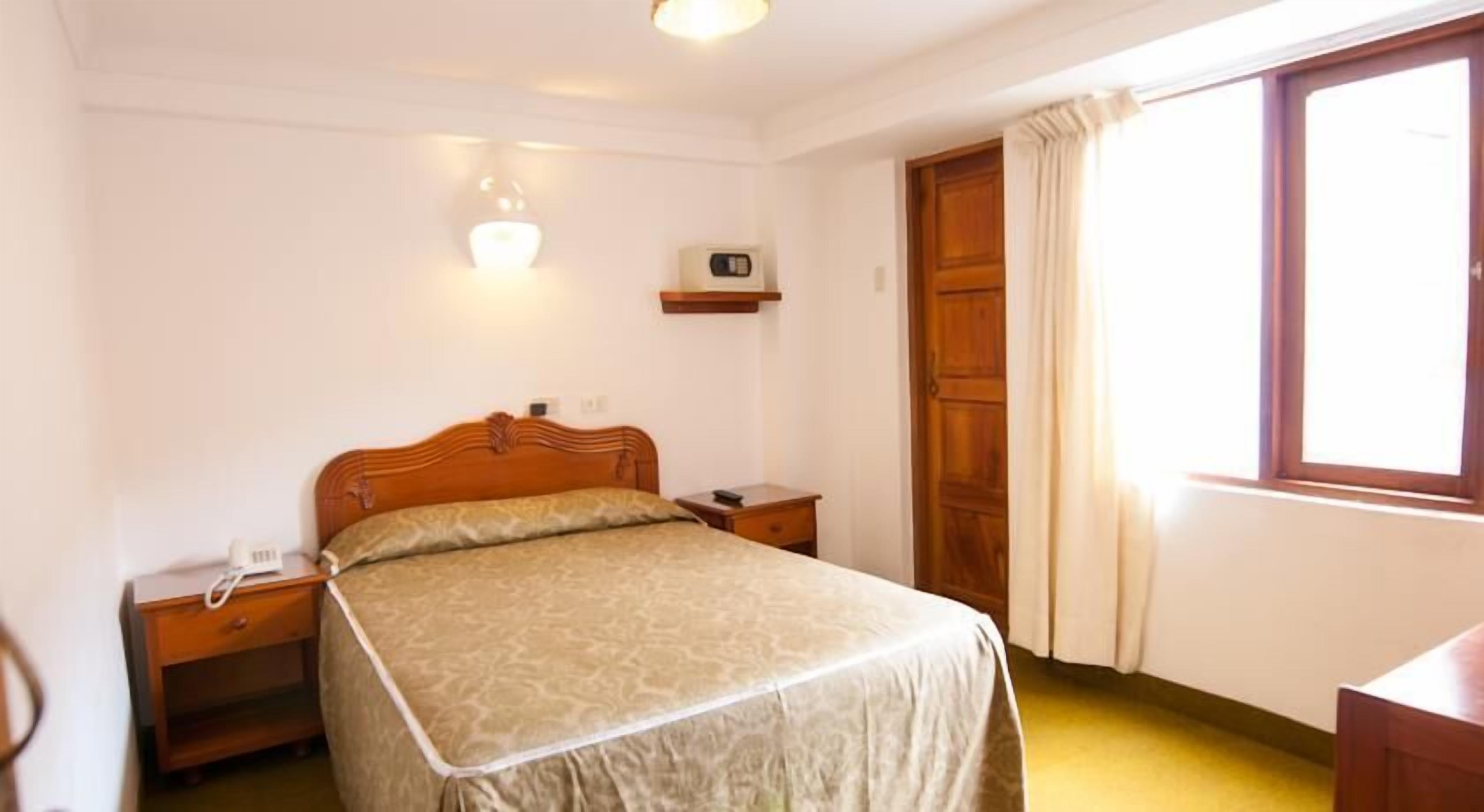 A bedroom with a bed, bedside tables, a nightstand, a wall-mounted radio, and a window with curtains.