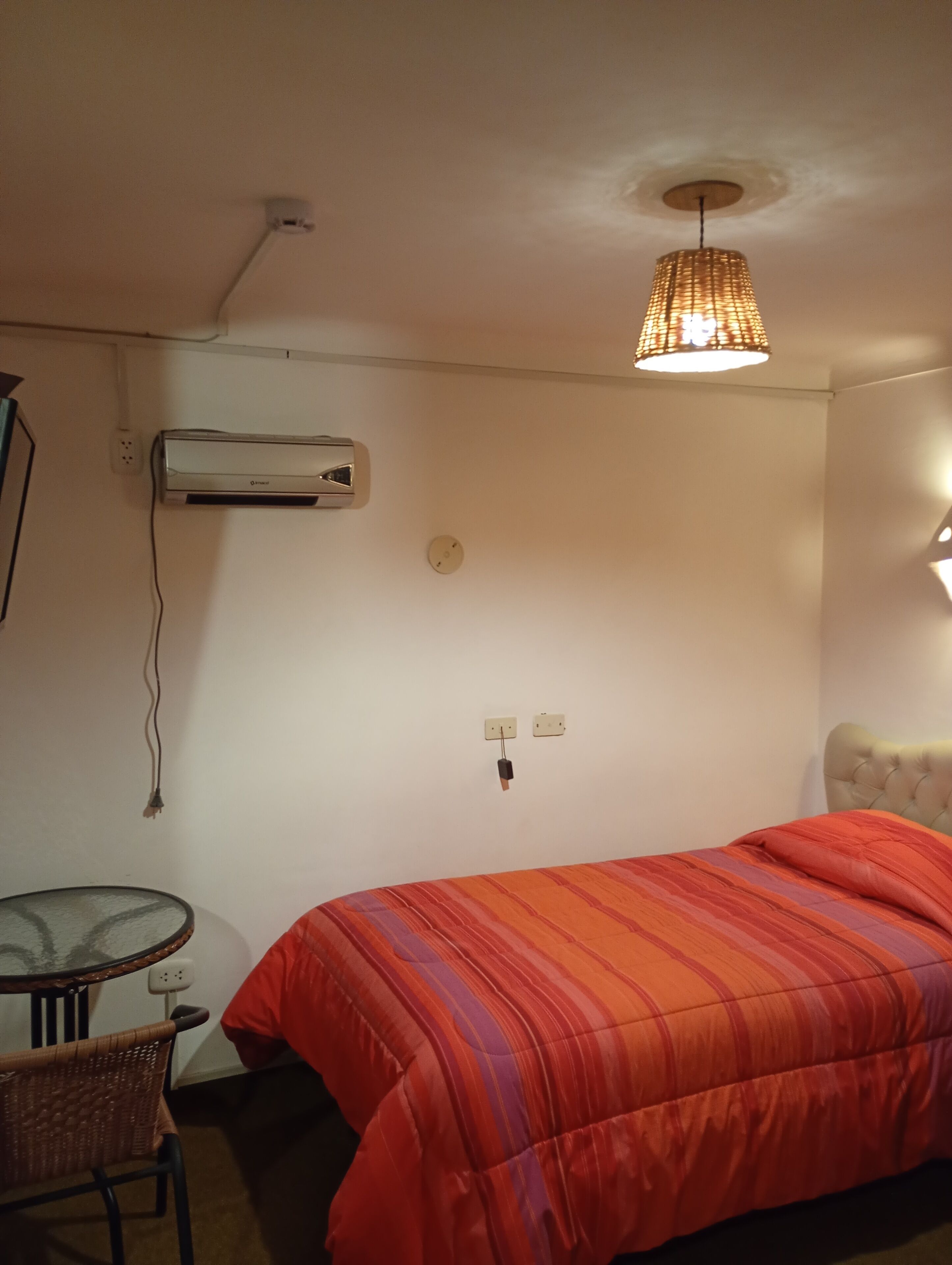 Een slaapkamer met een bed, een tafel, een stoel, een airconditioner en een hanglamp.