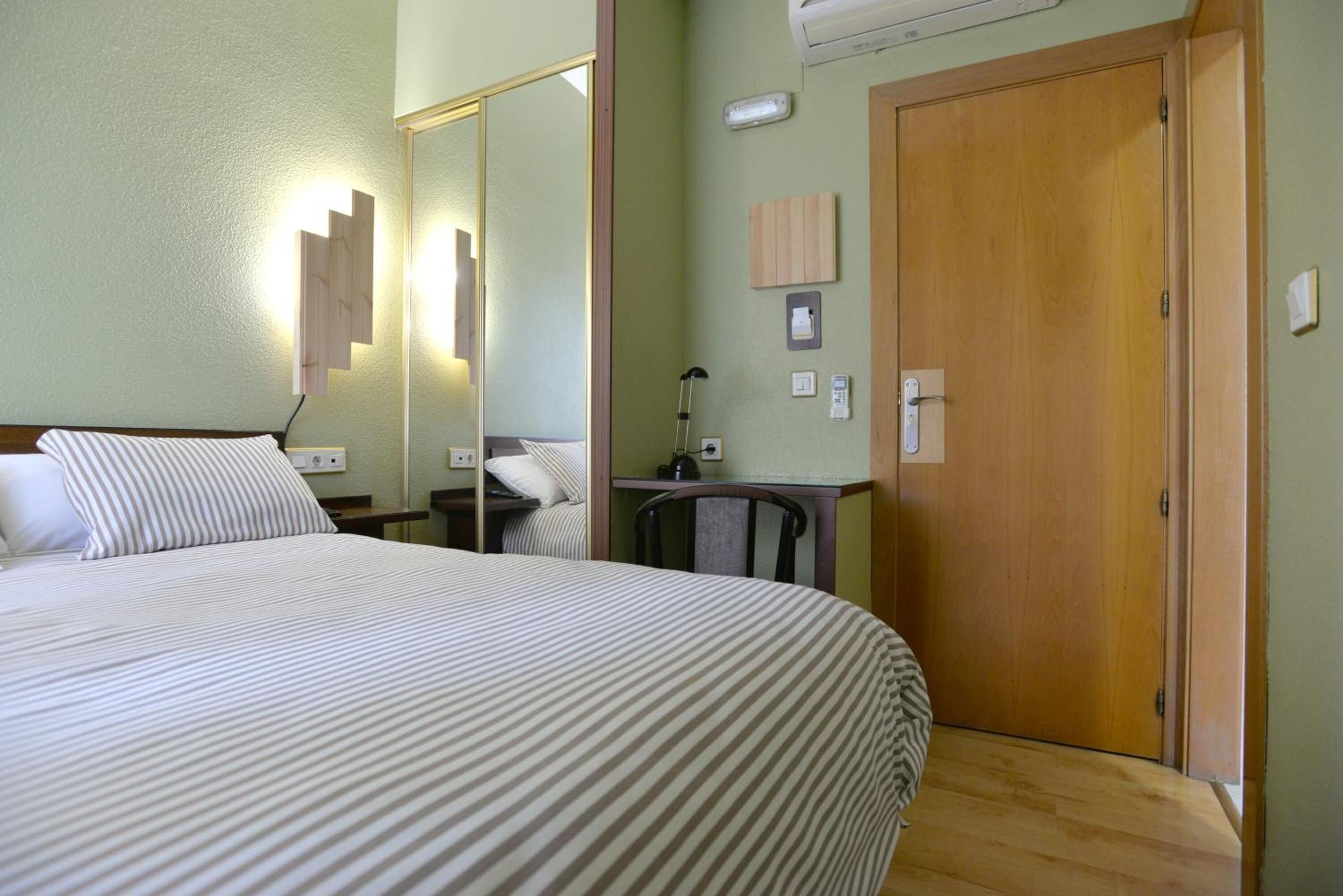 Habitación de hotel con una cama grande, una puerta de madera, un espejo, una mesita de noche y un pequeño escritorio con una silla.