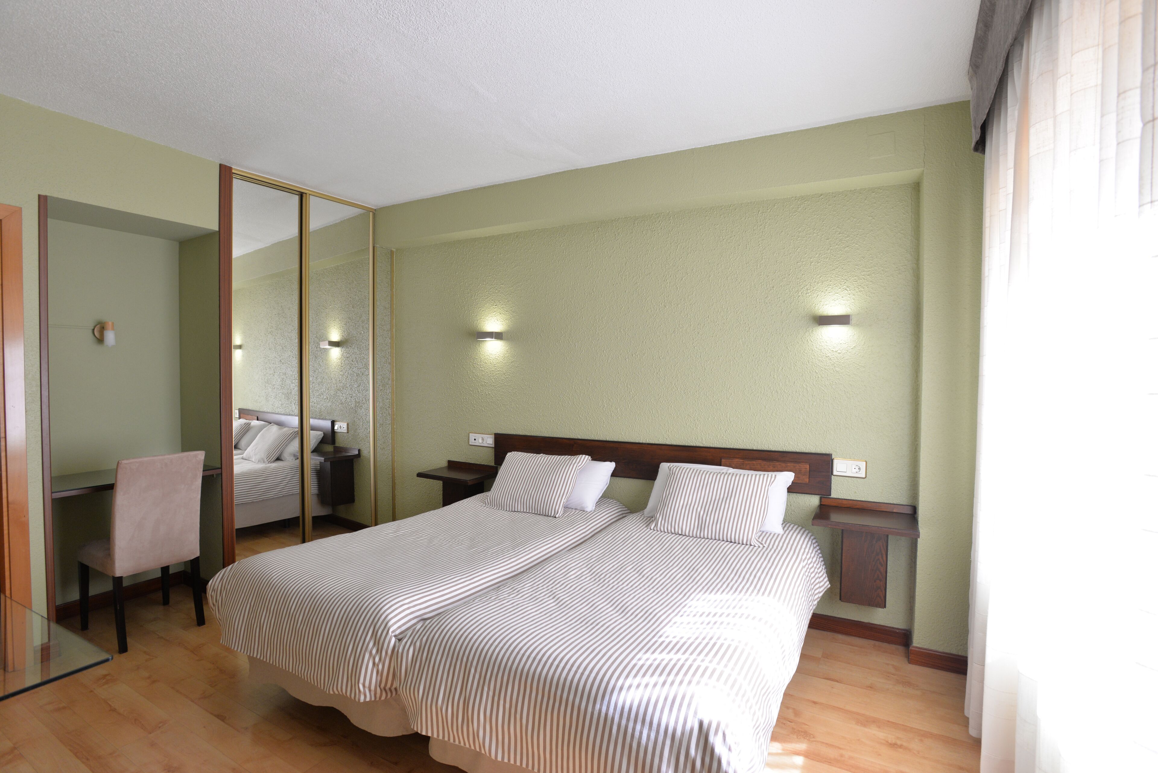 A hotel room with a large bed, a desk, a chair, and a mirror.