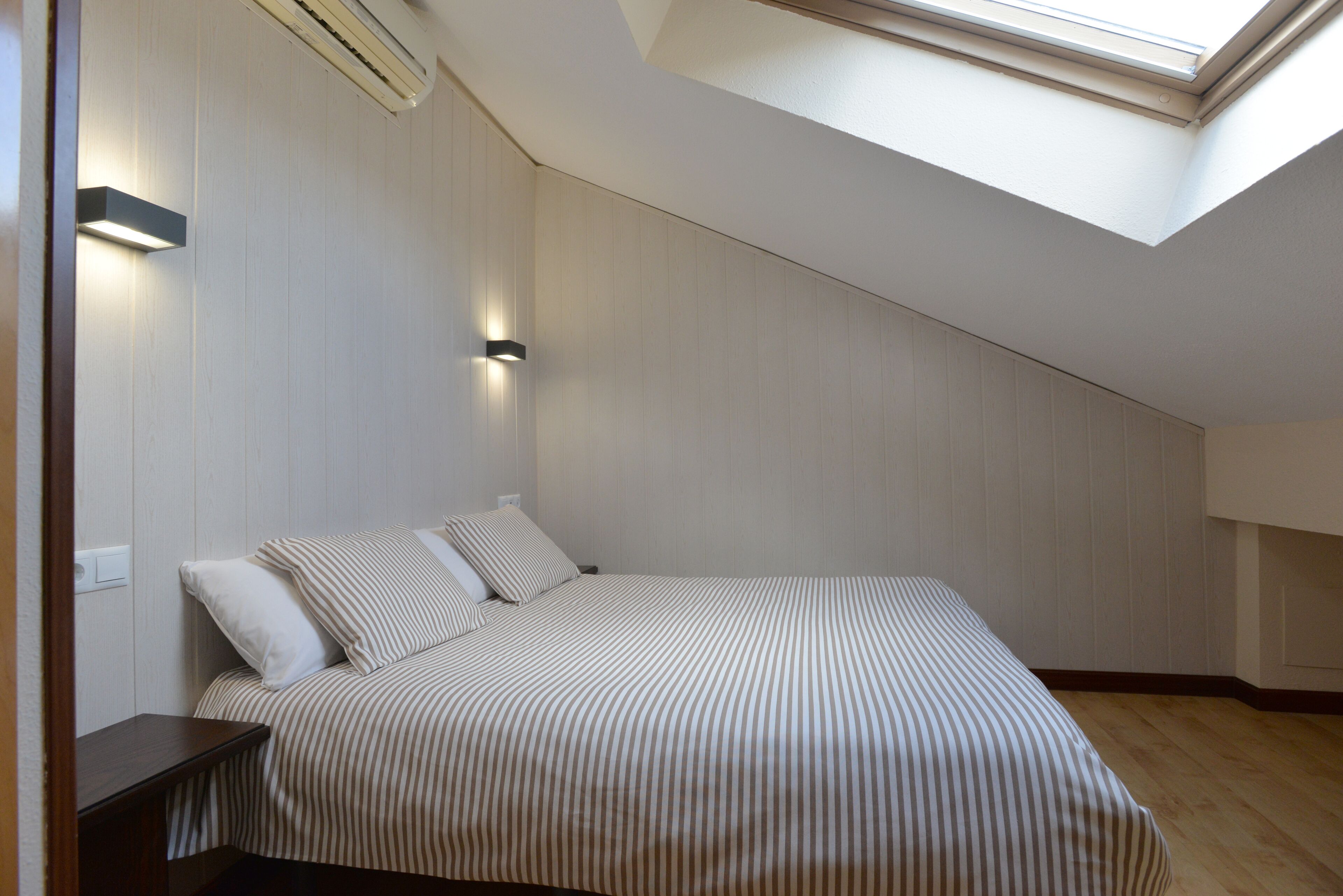 A bedroom with a sloped ceiling, a skylight, a bed with striped bedding, and two wall-mounted lights.
