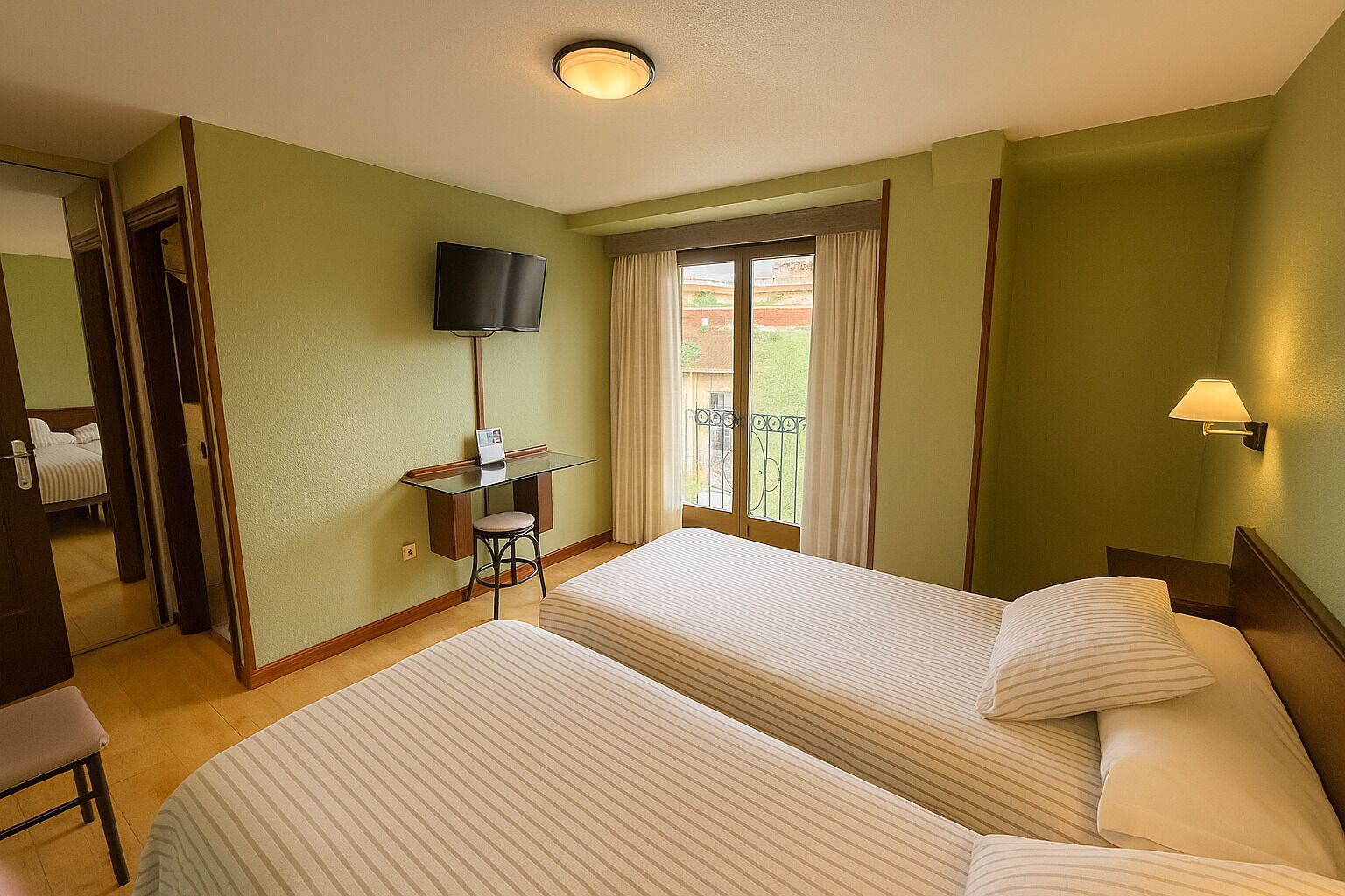 A hotel room with two beds, a desk with a chair, a television, and a balcony with curtains.