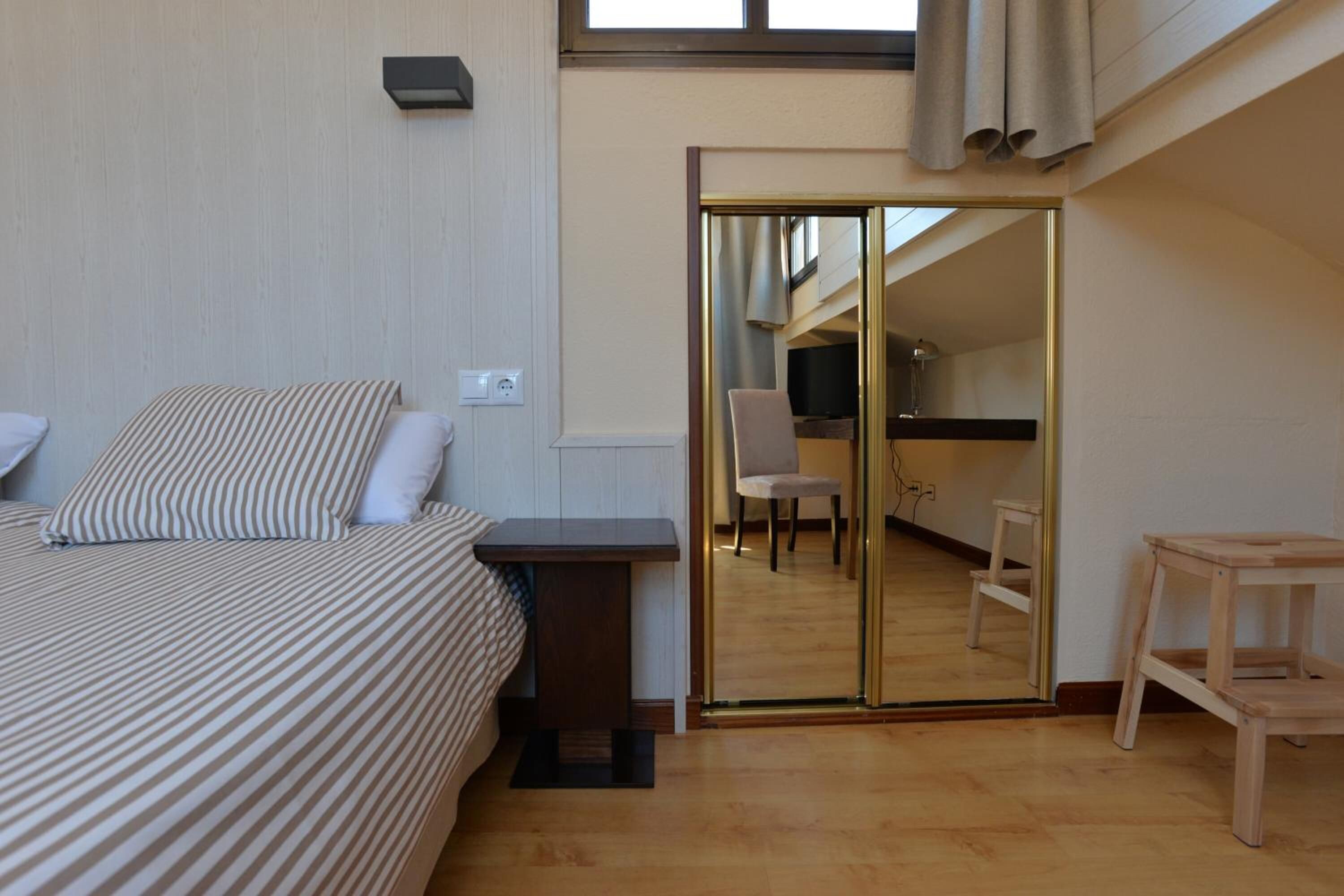 Un dormitorio con cama, una mesita, una silla y un espejo que refleja una cocina.