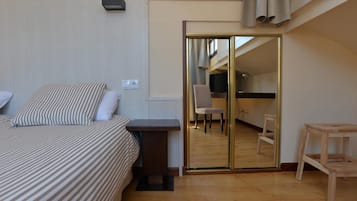 Un dormitorio con cama, una mesita, una silla y un espejo que refleja una cocina.