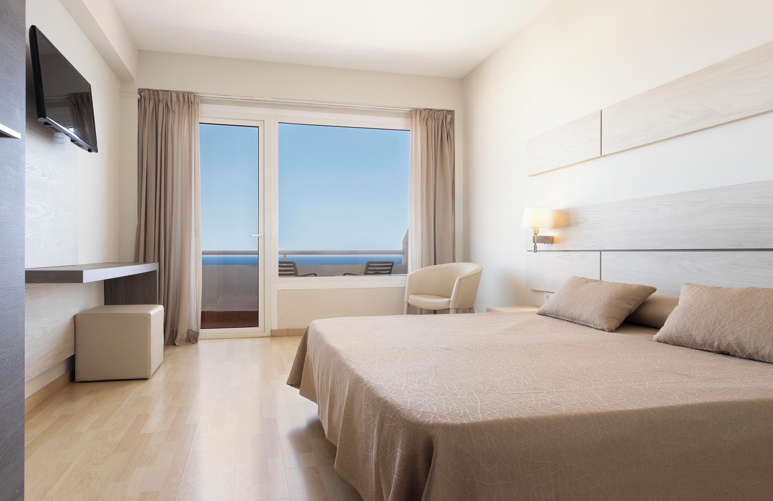 Habitació d'hotel amb un llit gran, una televisió penjada a la paret, una cadira i vista al mar a través d'una porta corredissa.