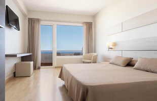 Habitación de hotel con una cama grande, un televisor montado en la pared, una silla y vistas al mar a través de una puerta corredera.