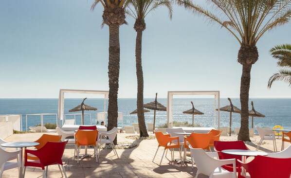 A beachside restaurant with outdoor seating, palm trees, and a view of the sea.