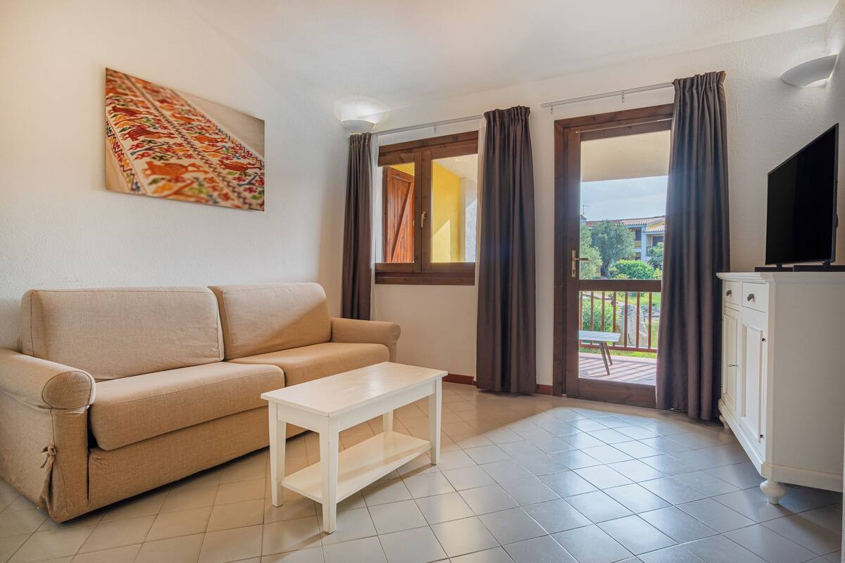 A living room with a beige sofa, a white coffee table, a TV, and a balcony with curtains.