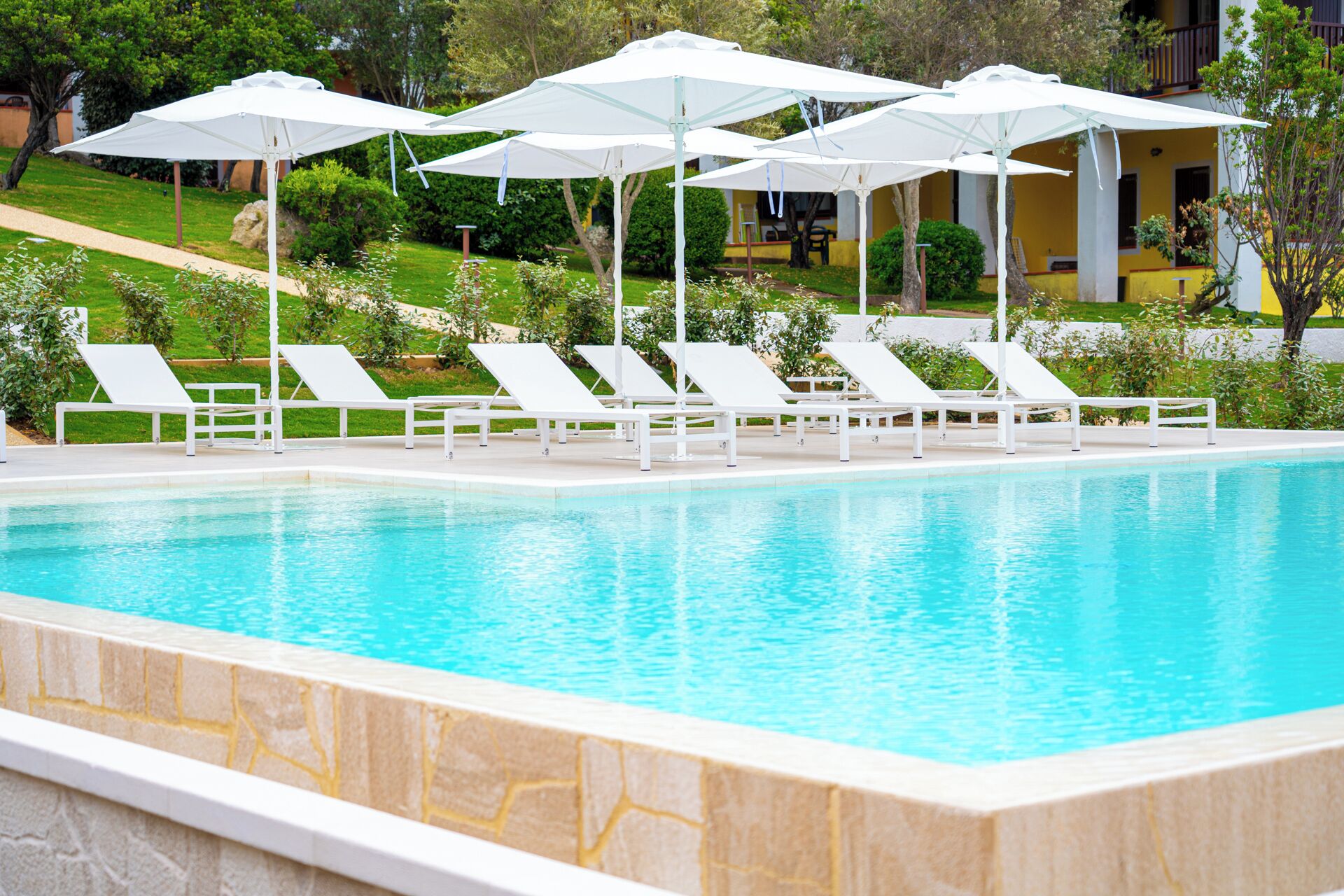 Una piscina con sillas de descanso blancas y sombrillas, rodeada de follaje y un edificio al fondo.
