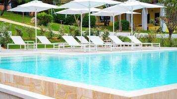 Una piscina con sillas de descanso blancas y sombrillas, rodeada de follaje y un edificio al fondo.