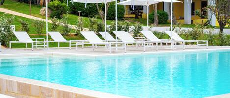 A swimming pool with white lounge chairs and umbrellas, surrounded by greenery and a building in the background.