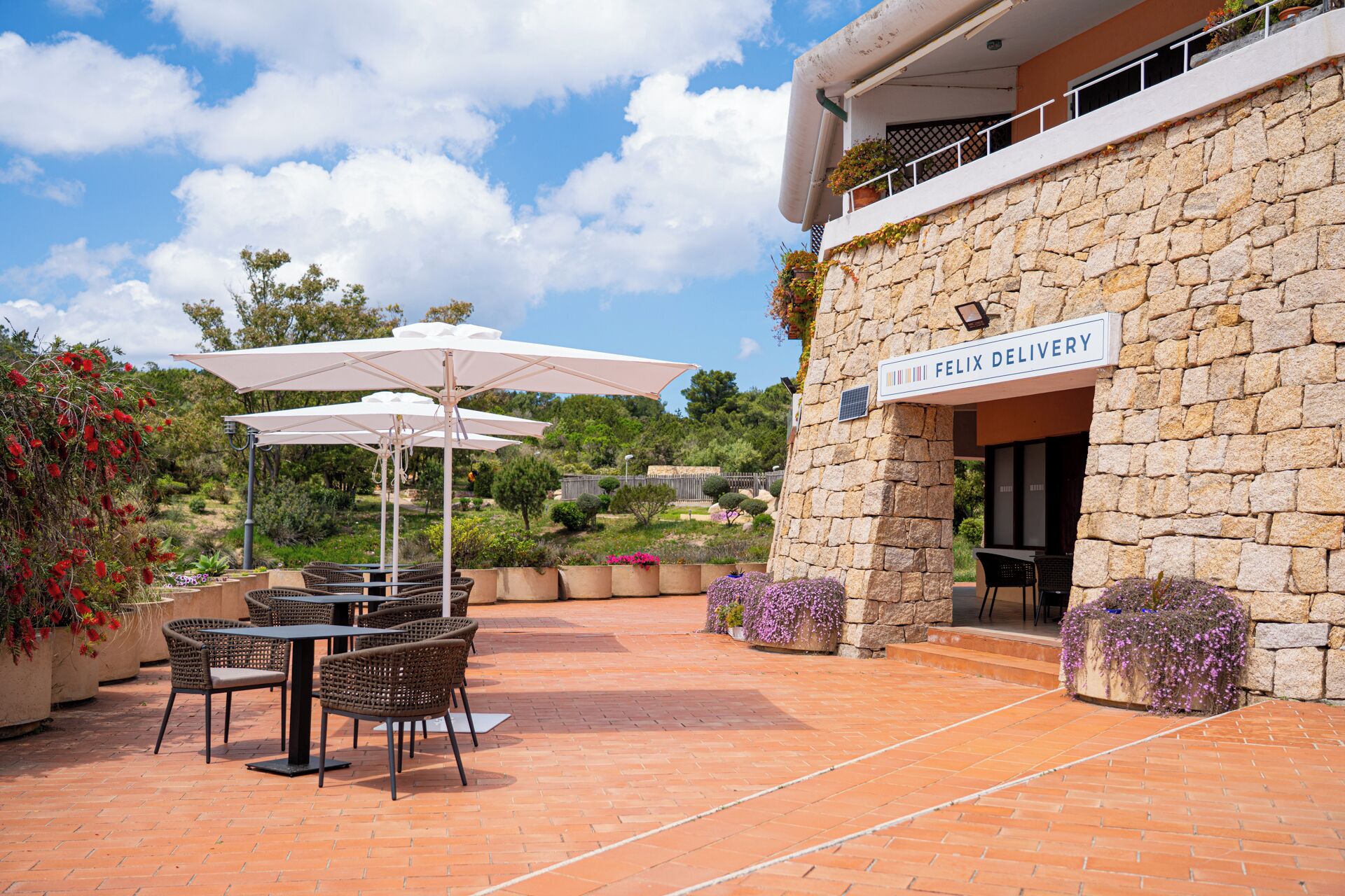 An outdoor patio with wicker furniture and white umbrellas, a stone building with a sign reading "FELIX DELIVERY", and a landscaped garden with various plants and flowers.