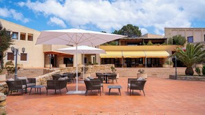 Un'area patio esterna con mobili in vimini e ombrelloni, un ristorante con tenda a righe gialle e un muro in pietra.