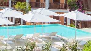 Area piscina con ombrelloni bianchi e poltrone prendisole, circondata da un edificio con balconi e piante.
