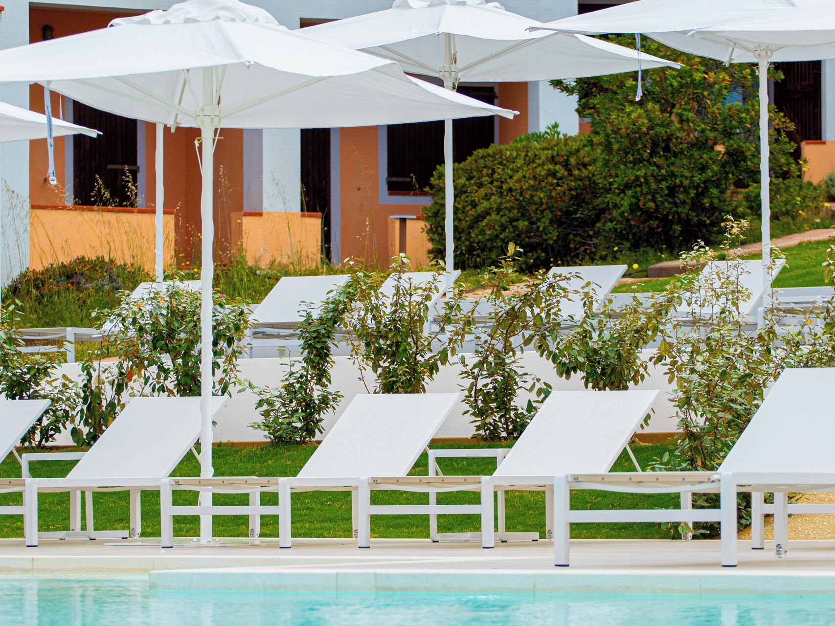 Un espace aménagé au bord de la piscine, avec des chaises longues blanches et des parasols, une piscine à l'eau claire, et un bâtiment aux murs orange, avec une haie verte en arrière-plan.