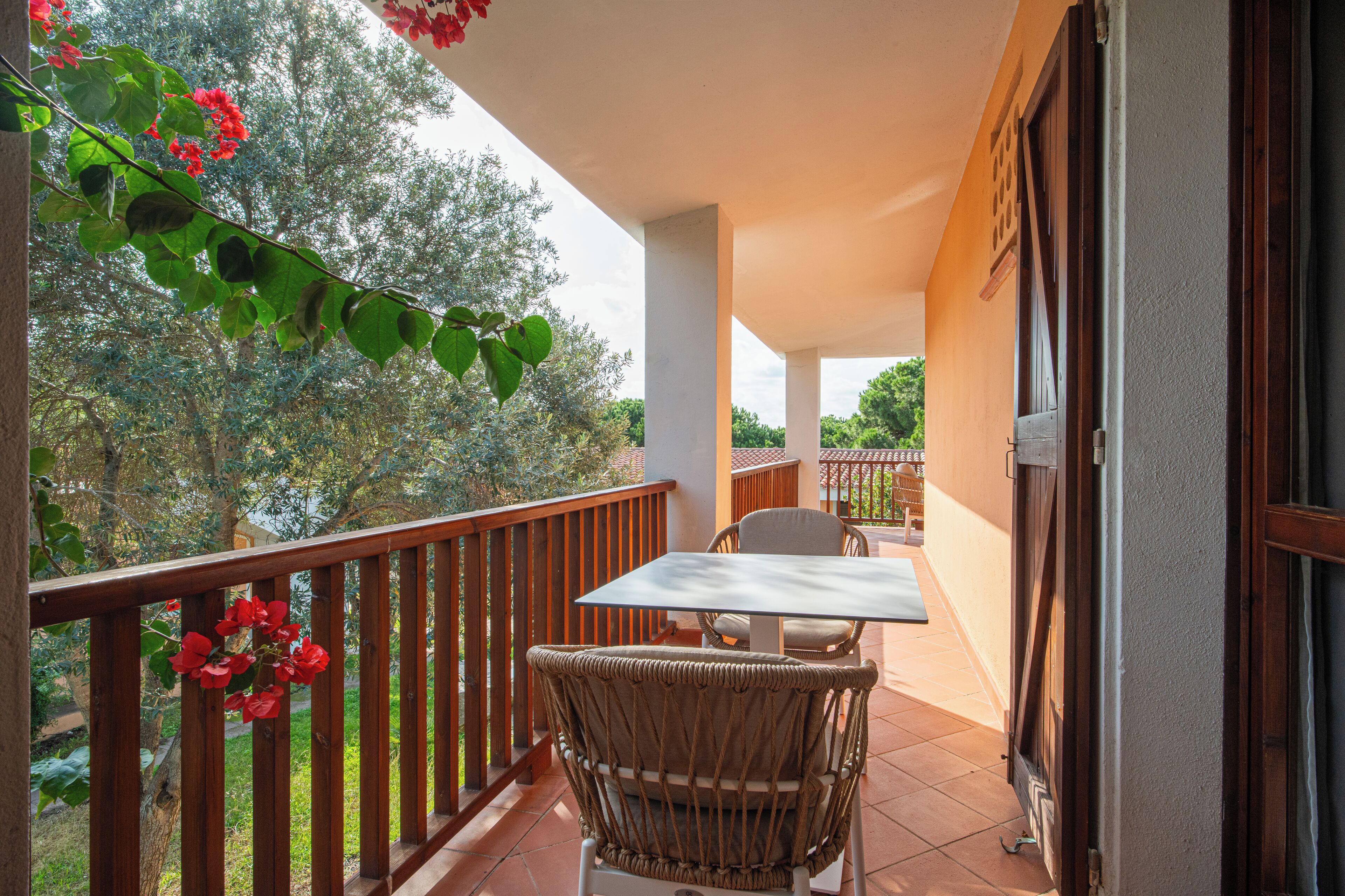 A balcony with a table and chairs, overlooking a garden with trees and red flowers.