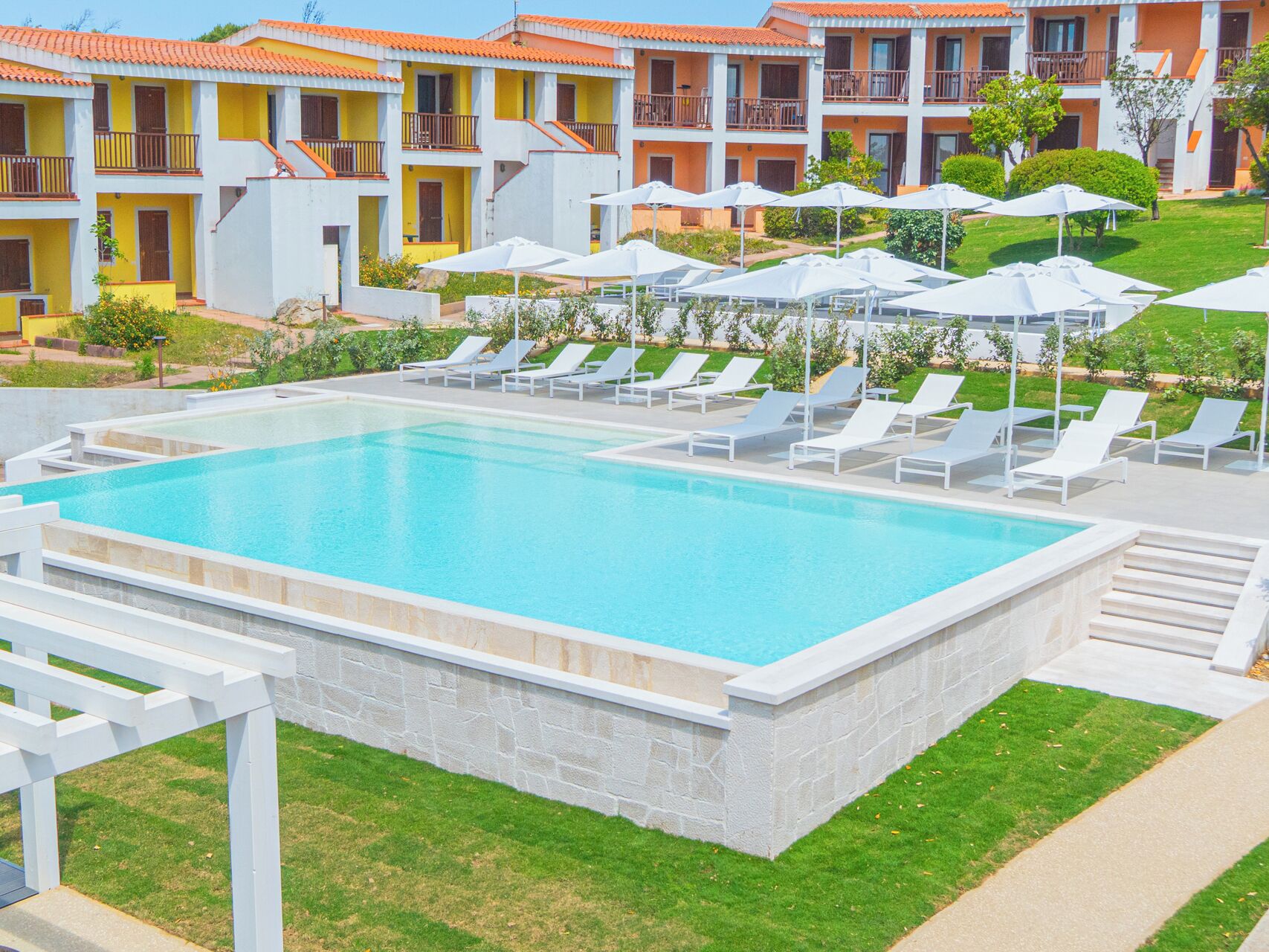 A swimming pool with white lounge chairs and umbrellas, surrounded by a well-maintained lawn and a multi-story building with balconies.