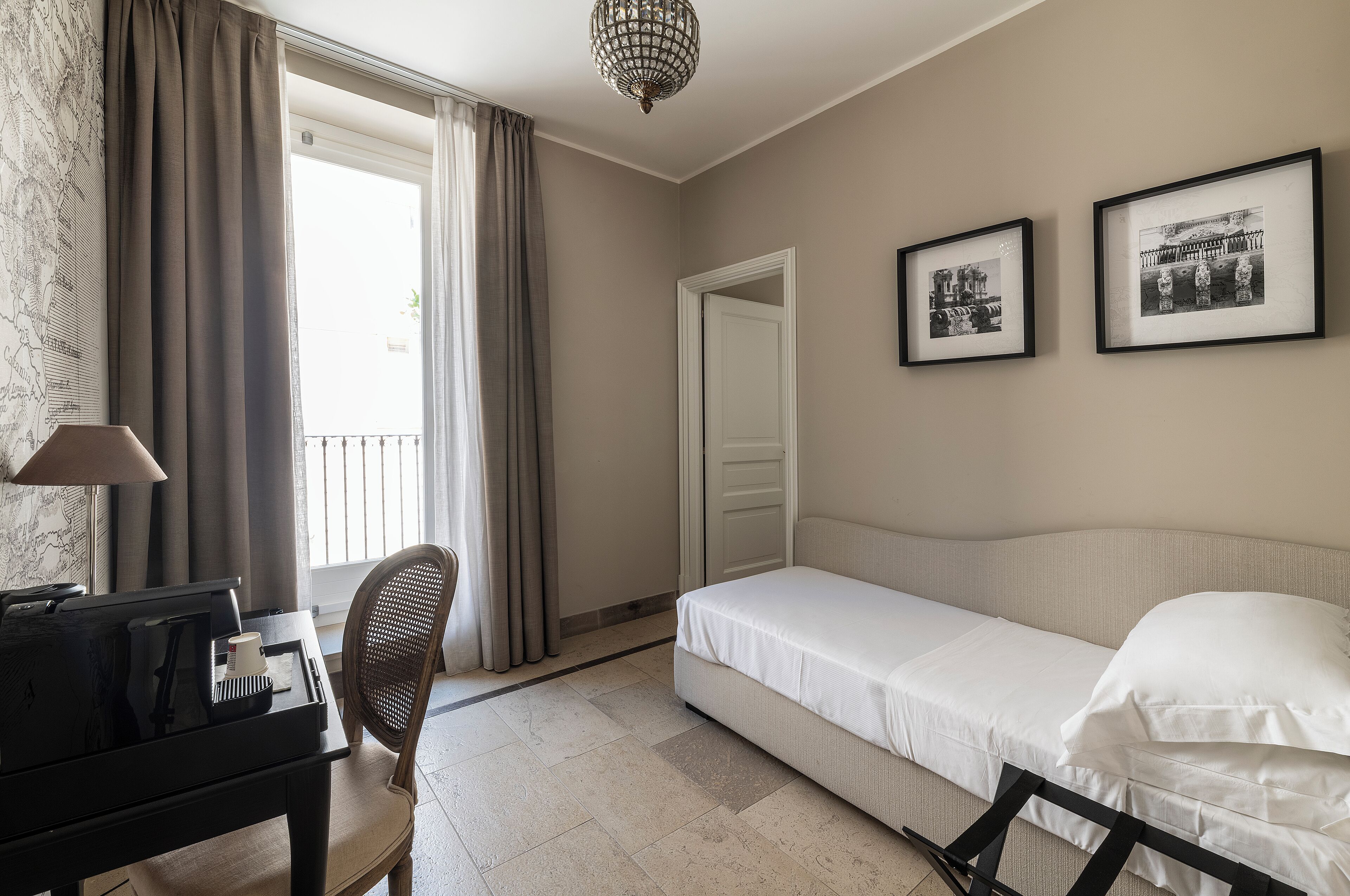 A hotel room with a bed, a desk with a lamp, a chair, and two framed pictures on the wall.