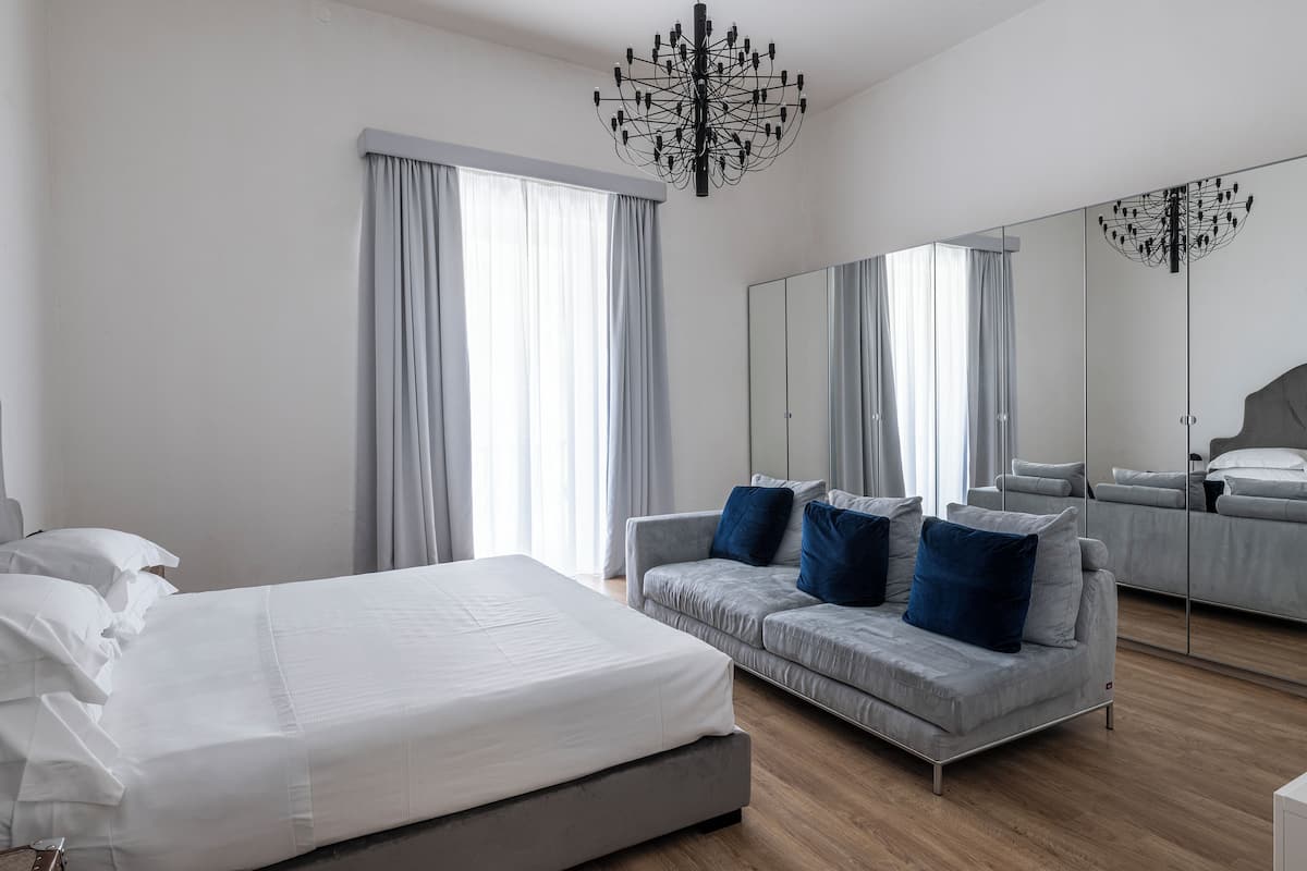 A modern hotel room with a large bed, a grey sofa with blue cushions, a large mirror, and a chandelier.
